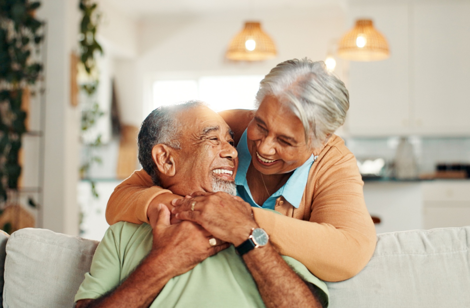 A smiling senior sits on a sofa as their partner embraces them from behind