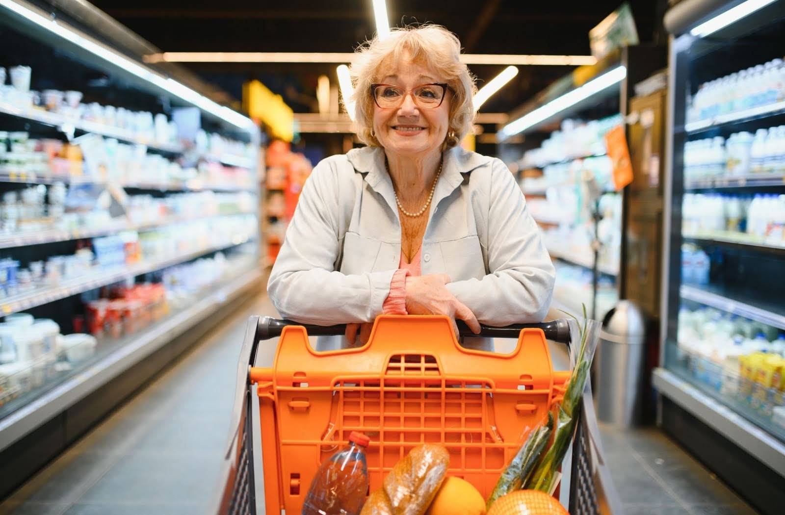 A senior smiles while pushing an orange shopping cart around a grocery store. 